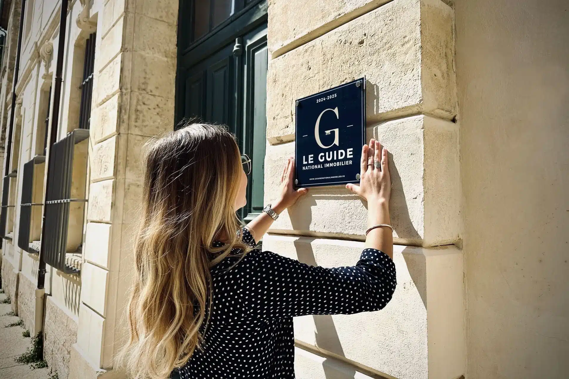 Une personne installe une plaque « Le Guide National Immobilier » sur la façade d’un bâtiment, illustrant la valorisation d’agences grâce à la création de site immobilier sur mesure.