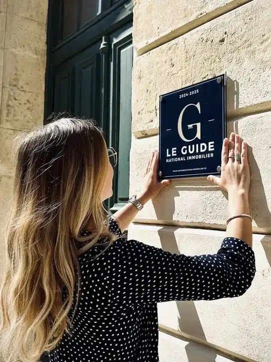 installation-plaque-immobiliere-gni Une personne fixe une plaque « Le Guide National Immobilier » sur la façade d’un bâtiment, illustrant la valorisation d’une agence grâce à une création site immobilier sur mesure et une identité professionnelle forte