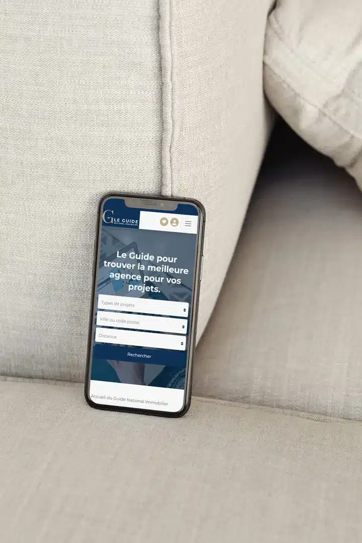 Un smartphone posé sur un canapé affiche l’interface du Guide National Immobilier, illustrant l’importance d’une création de site immobilier sur mesure pensée pour une navigation mobile fluide.