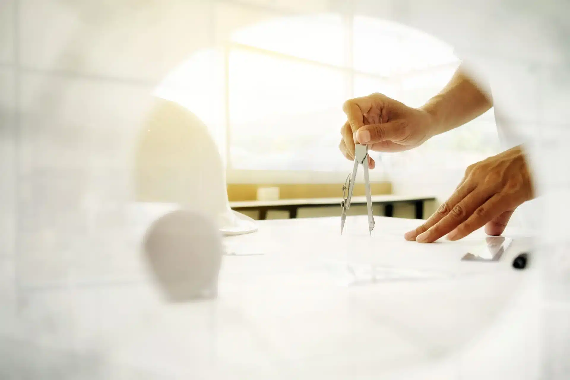 travail-dessin-sur-plan-avec-compas Main tenant un compas au-dessus d’un plan en cours de dessin, avec un casque de chantier en arrière-plan dans une pièce lumineuse, scène évoquant un travail de précision ; contenu utilisé dans un contexte lié à la création de plateforme de gestion d’adhérents